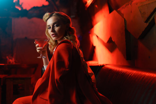 Image of young blonde in red jacket looking at camera with cocktail in her hand in nightclub