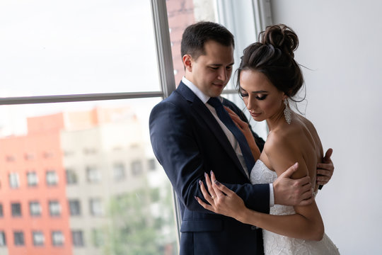 bride and groom hugging near window. feeling of love in newlyweds. guy and girl in wedding clothes pose in classical interior.