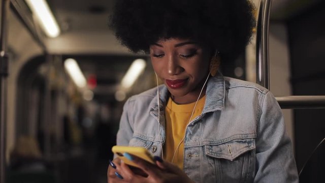 Portrait of young african american women in headphones listening to music and browsing on mobile phone in public transport. City lights background. Slow motion.