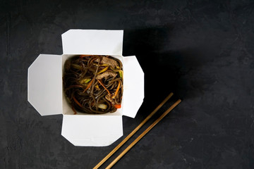 Asian restaurant food delivery. Soba noodles with meat, vegetables and soy sauce in white take-out paper box on black background with chopsticks, top view, copy space