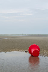 Bouée rouge posée sur le sable à marée basse sous un ciel gris. Côte belge
