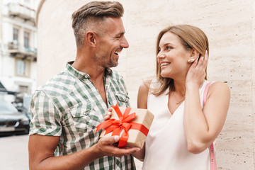 Image of romantic excited couple smiling and holding present box together while standing against wall on city street