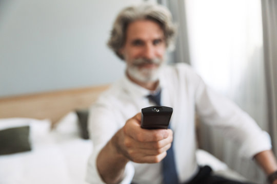 Image closeup of successful adult man holding remote control while watching television on bed in hotel apartment