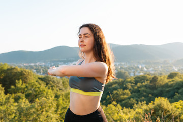 Naklejka na ściany i meble Young cute woman warming up during a workout in the Park. The concept of yoga, sports and meditation. In the background view of the settlement and the mountains