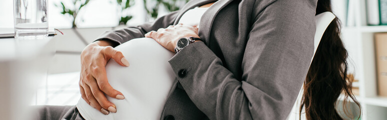 panoramic shot of pregnant woman holding belly while sitting in office