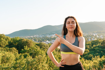 Naklejka na ściany i meble A young athletic woman points at something with her hand and looks at the camera. The concept of yoga, sports and meditation. In the background View of the village and the mountains. Copy space