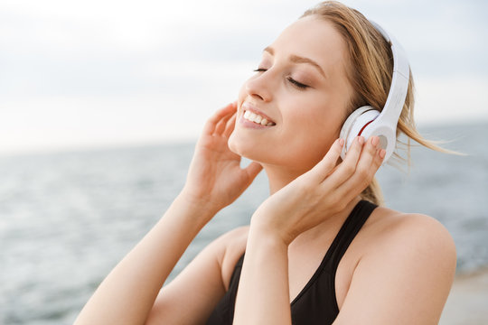 Image of happy beautiful woman listening to music with headphones near seaside in morning