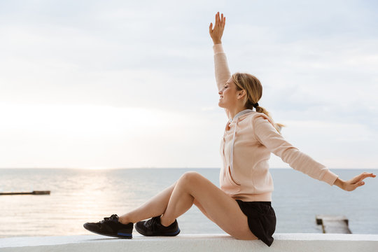 Image of caucasian woman smiling and throwing up arms while sitting near seaside in morning