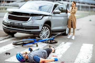 Road accident with injured cyclist lying on the pedestrian crossing near the broken bicycle and worried woman driver and car on the background