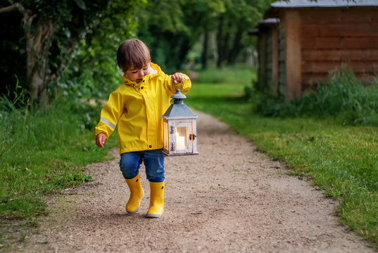 Little boy in yellow coat and rubber boots walking on path in dusk holding lantern with candle looking at light.