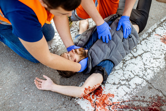 Medic with car driver applying first aid to the injured man lying on the pedestrian crossing after the road accident