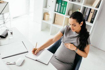 high angle view of pregnant woman taking notes in notebook while sitting behind table near bookcase