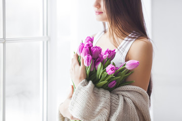 Attractive young woman with flowers indoors in the bedroom. Portrait of beautiful lady at home. Close up shot of female with tulips.