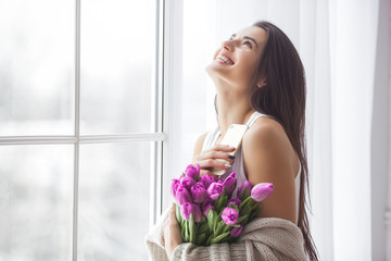 Attractive young woman with flowers indoors in the bedroom. Portrait of beautiful lady at home. Close up shot of female with tulips.