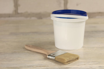 A paint brush is next to a plastic paint bucket with a blue lid on an old white vintage wooden board. Place for text or logo.