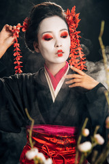 selective focus of beautiful geisha in black kimono with red flowers in hair and sakura branches on black background with smoke
