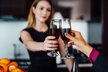 Young women wearing elegant black dress holding a bottle of red wine and a glass standing at kitchen bar looking at her female friend