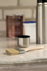 A brush lies next to a plastic paint bucket with a blue lid, a metal can, a thermos with a cup on an old white vintage wooden board. Place for text or logo.