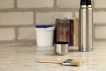 A brush lies next to a plastic paint bucket with a blue lid, a metal can, a thermos with a cup on an old white vintage wooden board. Place for text or logo.