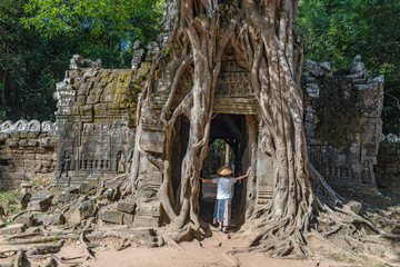Naklejka premium One tourist visiting Angkor ruins amid jungle, Angkor Wat temple complex, travel destination Cambodia. Woman with traditional hat, rear view.