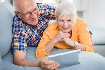 Happy senior couple looking at tablet lying on sofa at home