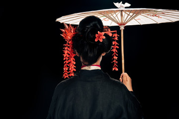 back view of beautiful geisha in black kimono with red flowers in hair holding traditional asian...