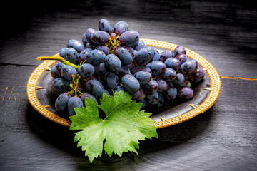 Bunch of ripe blue-black table grape with leaf served on black plate on black wooden background