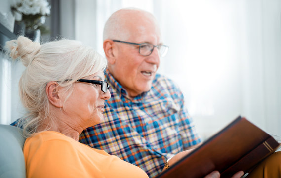 Cheerful senior couple holding family photo album sitting on sofa at home