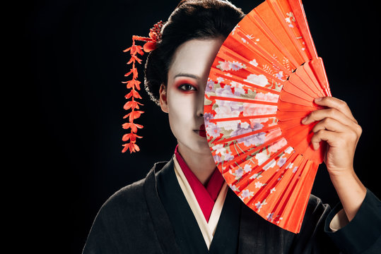 attractive geisha in black kimono with flowers in hair holding bright hand fan isolated on black