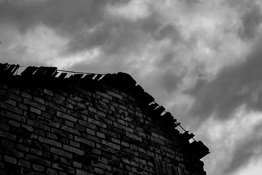 Black and white image of a brick building with a leaky roof on a cloudy day. Old barn against a rainy sky.
