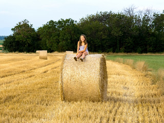 Naklejka na ściany i meble The little girl sits on a haystack in the twilight of day