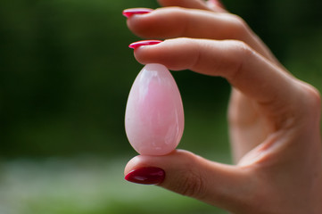 Close up photo of female hand holding a yoni egg. Rose quartz crystal egg on river background. Womens health concept