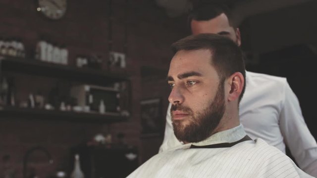 Serious young male hairdresser is cutting beard and hair to regular customer