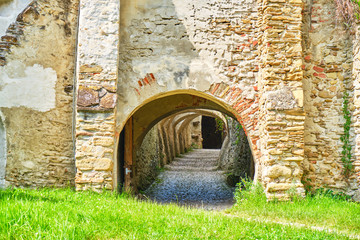 Tunnel with multiple arches and cobblestone going through a brick stone wall, from the green grass outside to the interior of Biertan Fortified Church, in Transylvania, Romania.