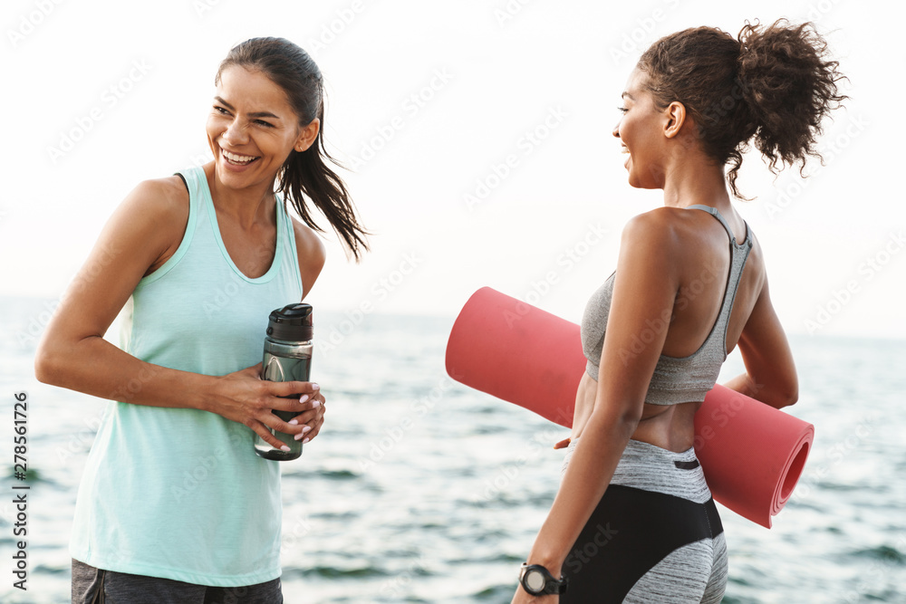 Sticker photo of two cheerful multiethnic sportswomen talking and laughing while standing with yoga mat by s