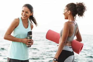 Photo of two cheerful multiethnic sportswomen talking and laughing while standing with yoga mat by seaside in morning