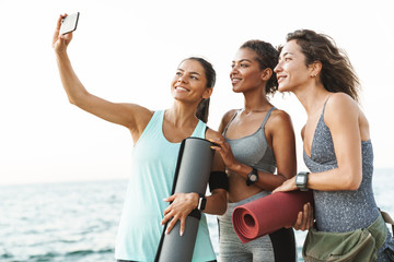 Photo of three feminine multiethnic sportswomen smiling and taking selfie on cellphone while standing by seaside in morning