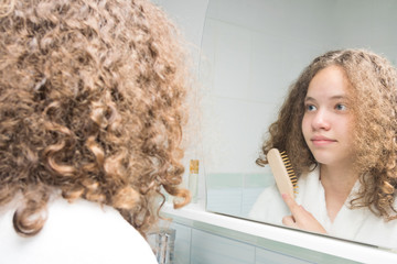 girl in a white bathrobe in the bathroom standing in front of the mirror combing her curly hair