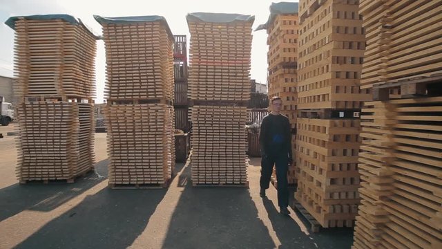 A young worker in uniform walks between wooden pallets in distribution outdoor warehouse. Slow motion