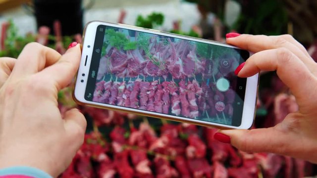 A woman photographs on the smartphone rows of delicious raw lamb for a barbecue or grill. Social network blogger concept photo. Taking picture using cell phne.