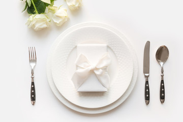 White table setting with bouquet rose, gift, dishware, silverware on white table. Romantic and wedding anniversary.