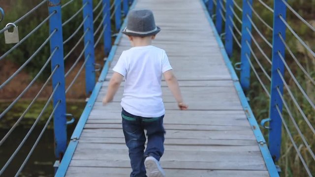beautiful toddler in t-shirt hat and jeans runs along blue wooden suspension bridge over river close backside view