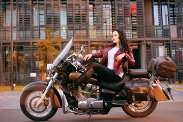 Fototapeta na wymiar A beautiful brunette is sitting on a bike near the office. The girl came to work on a motorcycle. Success. Girl Lezhin on a motorcycle in a leather jacket. Girl on a big black motorcycle.