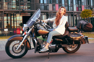 Fototapeta na wymiar Beautiful blonde sits on a motorcycle near the office. Girl came to work on a motorcycle. Success. Girl lies on a motorcycle in a jacket. Girl on a large black motorcycle on an industrial background.