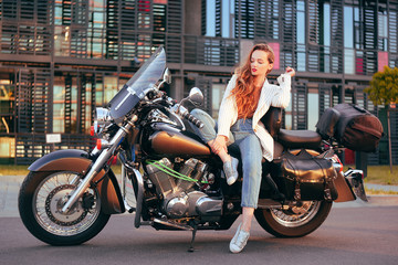 Plakat Beautiful blonde sits on a motorcycle near the office. Girl came to work on a motorcycle. Success. Girl lies on a motorcycle in a jacket. Girl on a large black motorcycle on an industrial background.