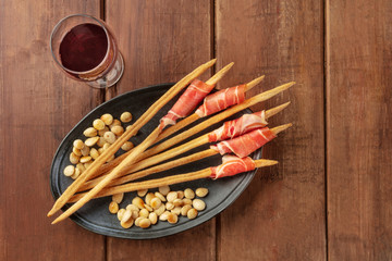Prosciutto-wrapped Italian grissini with wine and roasted almonds, top view. Italian antipasti with parma ham, shot from the top on a dark rustic wooden background with a place for text