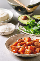 Ingredients for cooking poke bowl. Rice, soy sauce marinated salmon, avocado, tofu cheese and green salad in ceramic bowls served with wooden chopsticks over grey spotted table.