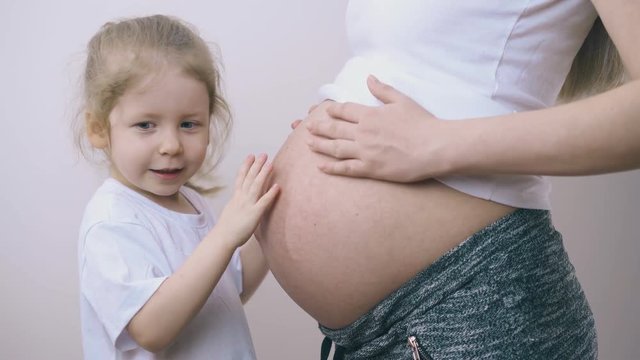 happy curly haired little girl plays with pregnant mother large belly standing near white wall at home