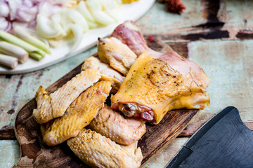 Pieces of fresh raw chicken parts on a cutting board on wooden old plank or table with blur Thai herbs, whole chicken and black knife. Chicken with Thai herbs prepared for cooking soup. Top view.