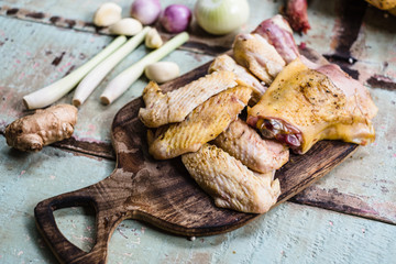 Pieces of fresh raw chicken parts on a cutting board on wooden old plank or table with blur Thai herbs and whole chicken. Chicken with Thai herbs prepared for cooking soup.